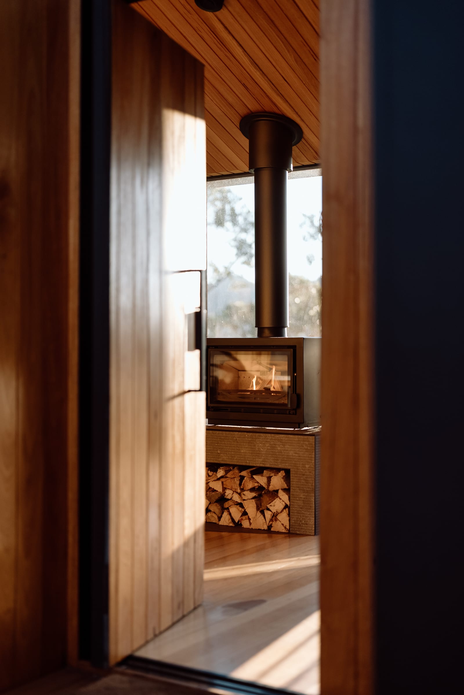 The view through a Hunter Huon Valley cabin's timber doorway to the wood fire alight, split logs stacked in the hearth below it and lined timber overhead