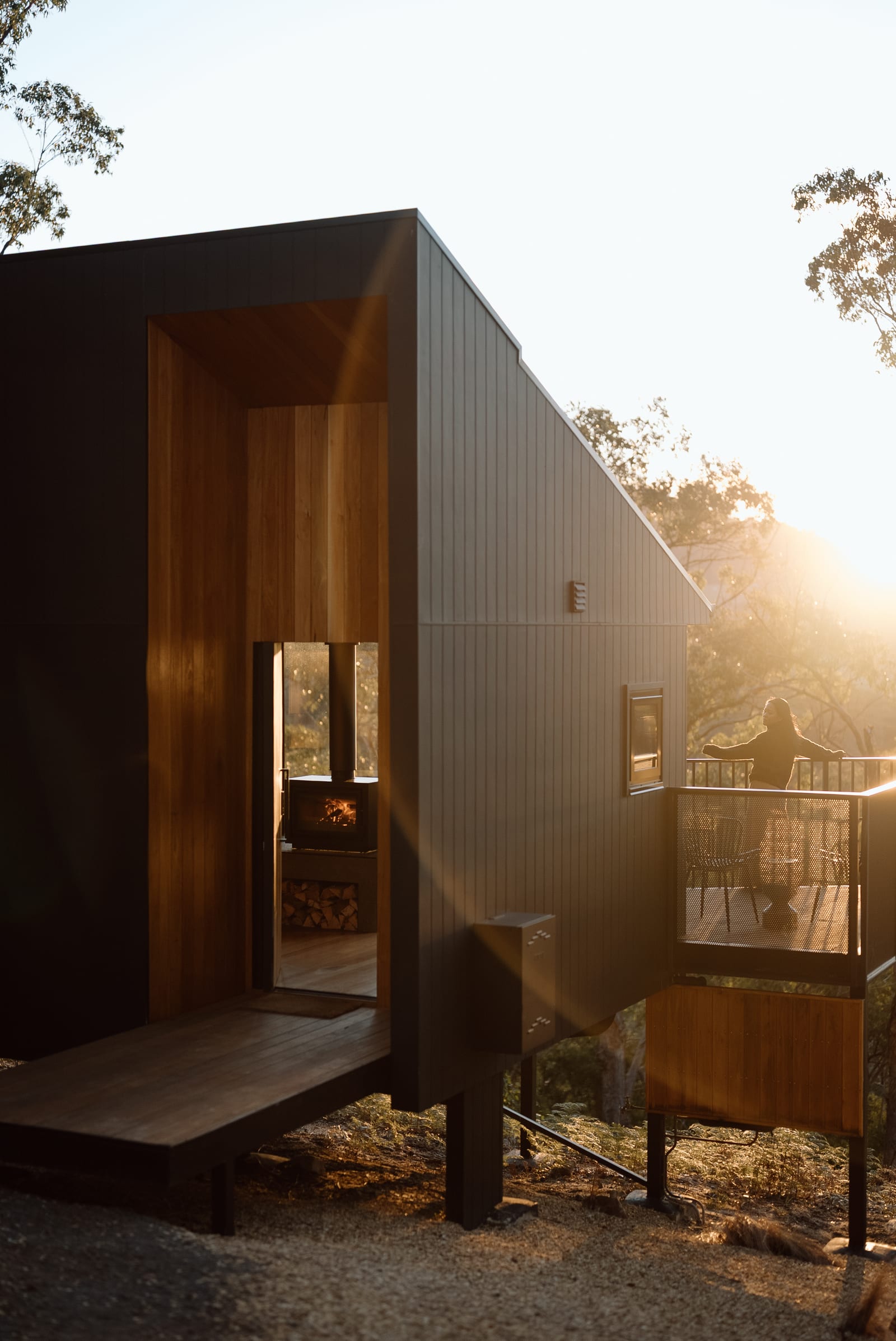 A black timber cabin at Hunter Huon Valley in low afternoon sun, its warm timber entry open to the deck, where someone stands at the rail looking out over the Glen Huon bush
