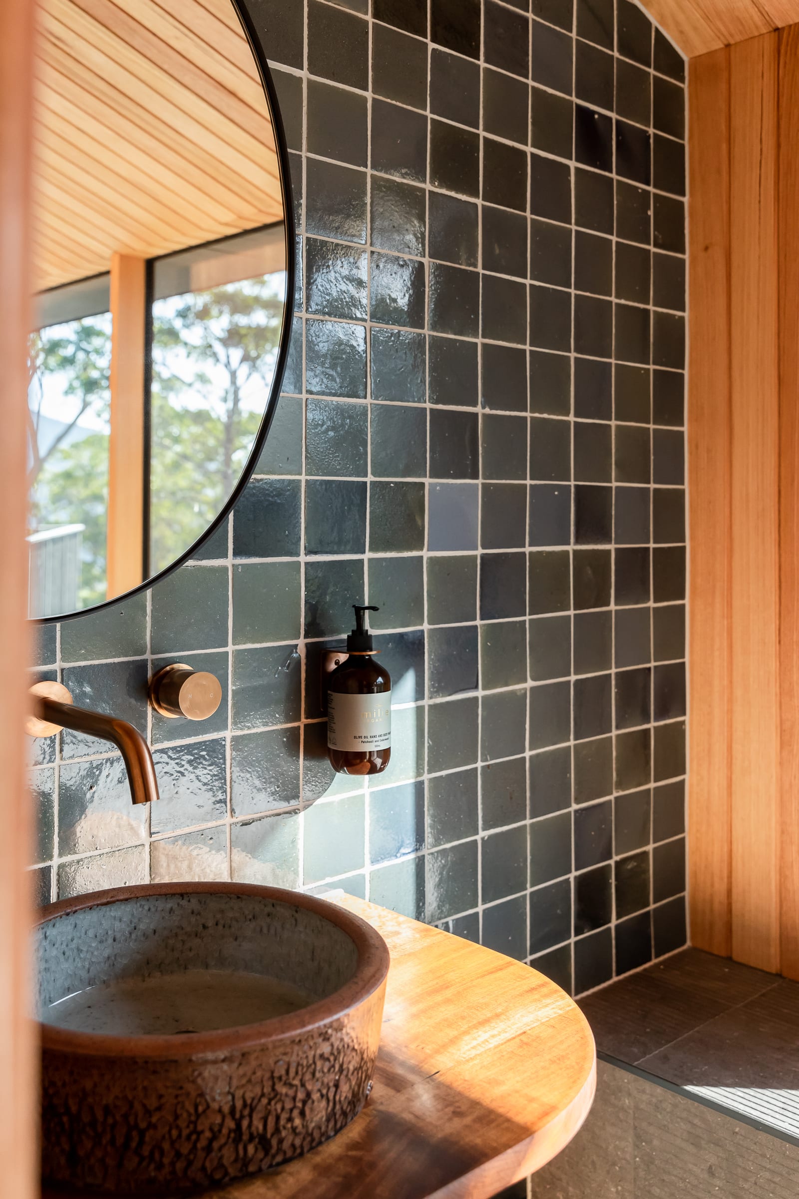 A textured stone basin on a timber vanity beneath a round black-framed mirror, with a copper tap, a single amber soap bottle and a wall of glossy dark green tile.