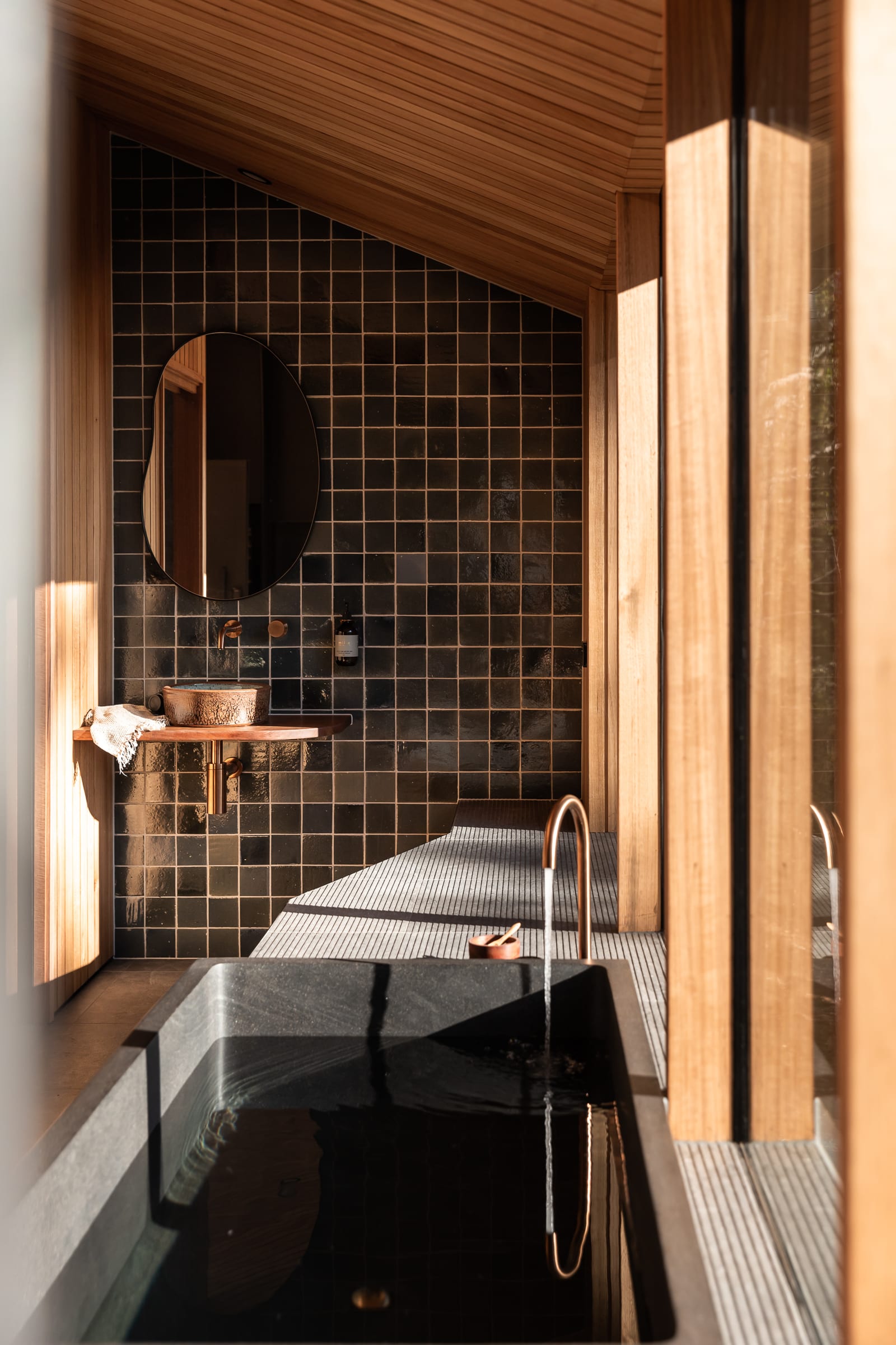 Looking down the length of the cabin past a filled stone bath to the ensuite, where a round mirror and a hammered basin on a timber shelf sit against a wall of dark green square tile.