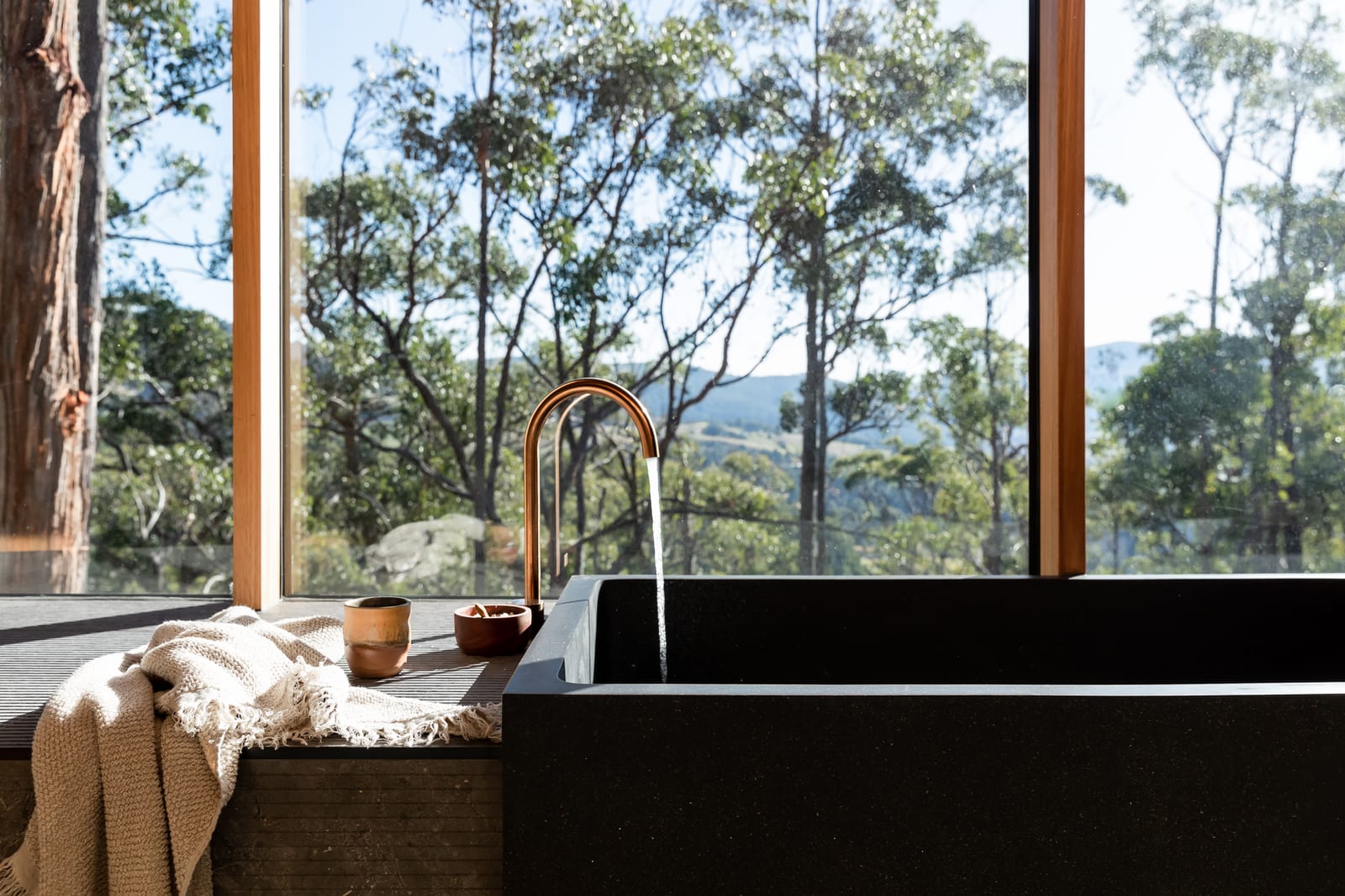 A copper tap arcing water into a dark stone bath set against full-height windows, a knit throw and two small ceramic cups on the ledge, with gum trees and a hazy ridge line beyond the glass.