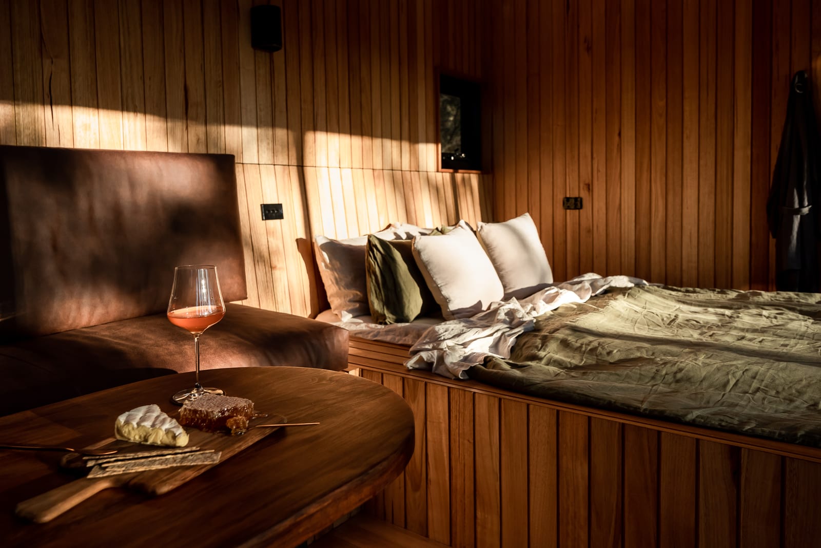 A glass of rosé and a board of cheese and honeycomb on a round timber table in the evening, with the leather bedhead and the olive-linen bed behind.