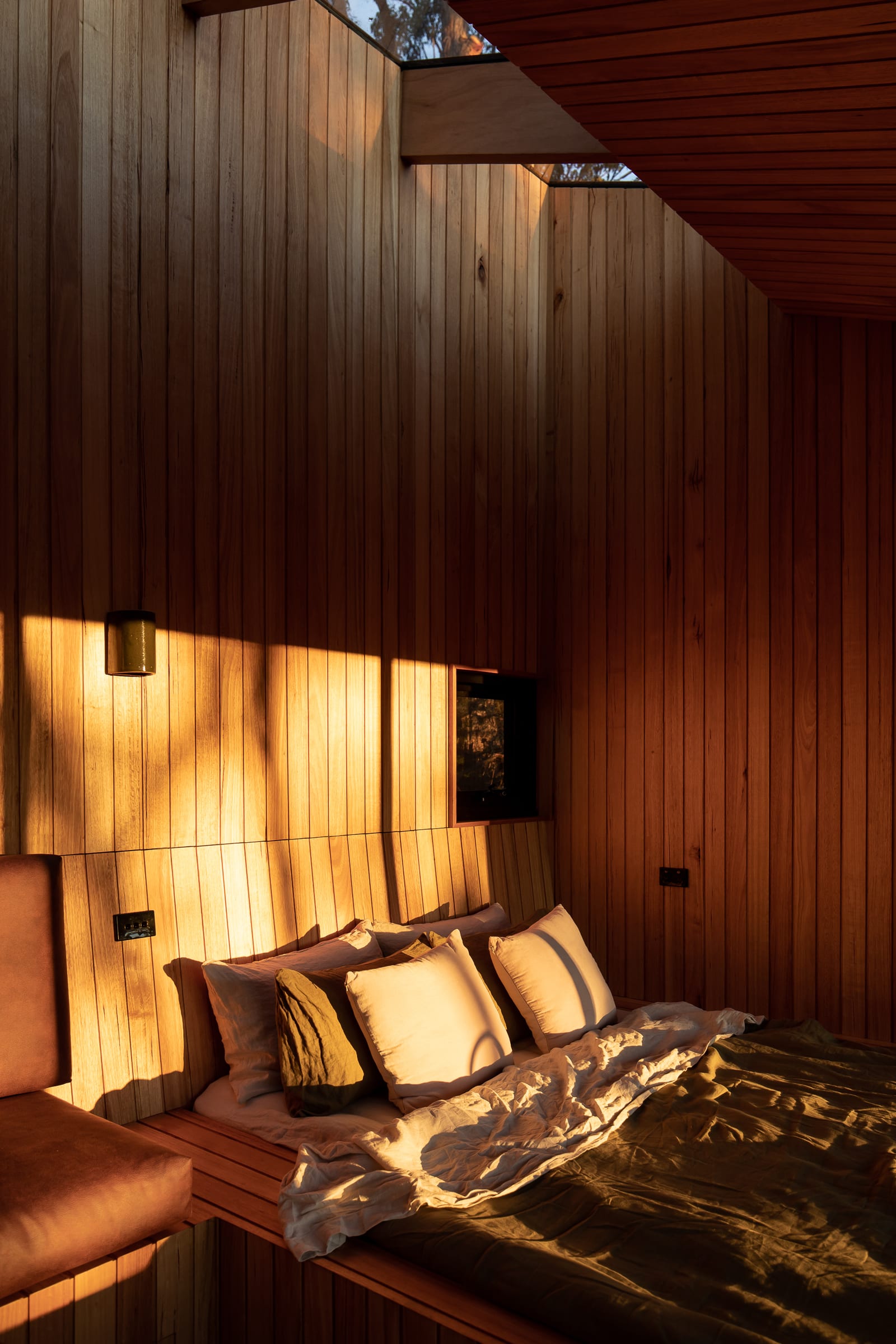 Late sun falling across a timber-lined bedroom wall in a hard diagonal, the bed made up in olive linen with layered pillows, and a skylight cut into the ceiling above.