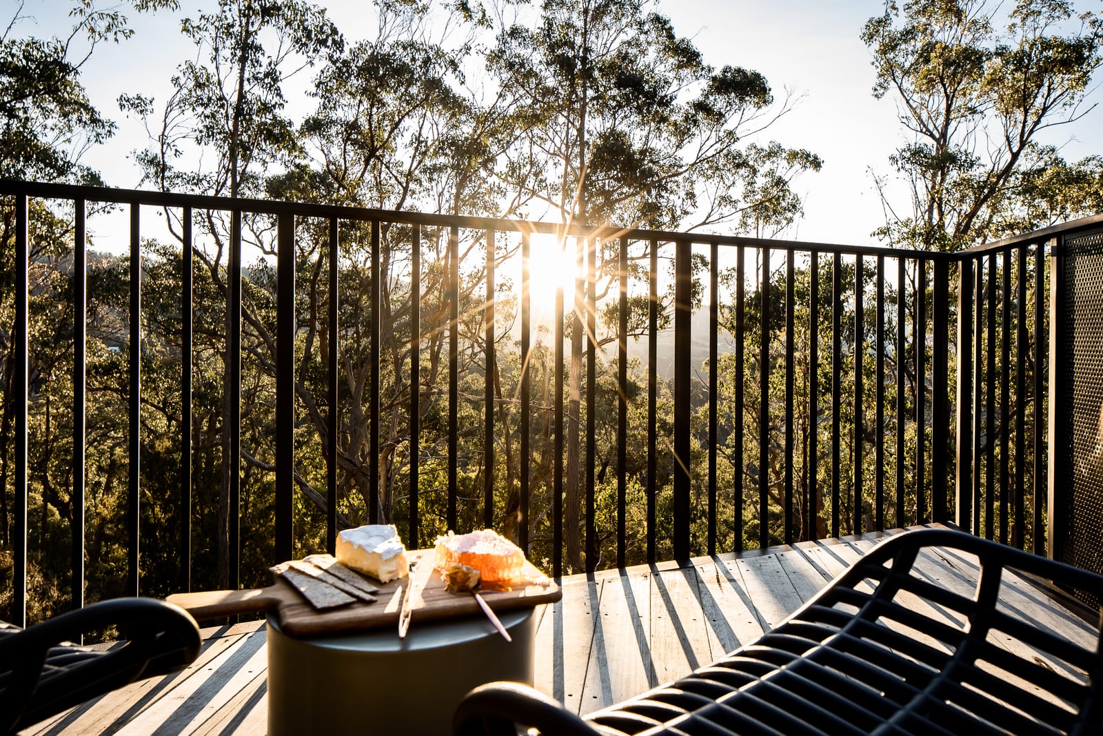 A board of cheese and fruit on a side table on the deck at sunset, the sun dropping through the trees behind the black balustrade and a wire lounger to the right.