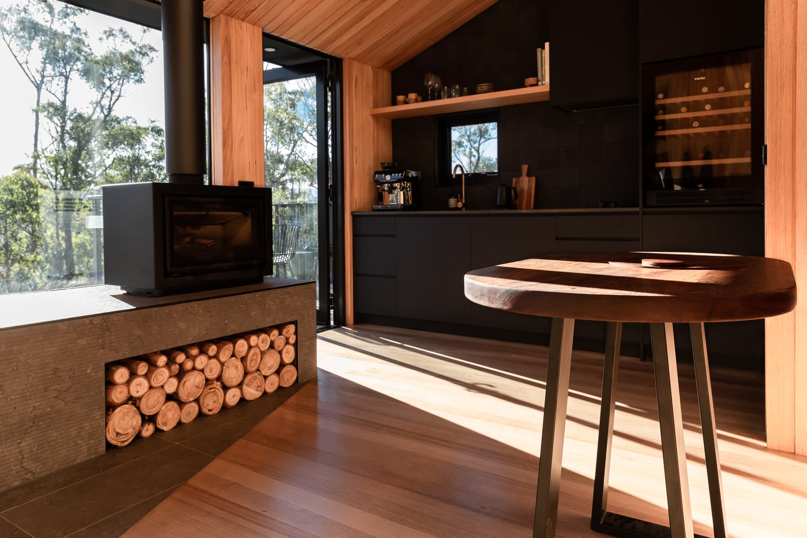 A wood fire burning in a black steel firebox above a recess stacked with split log ends, a live-edge timber table in the foreground and the black kitchen beyond, with the deck door open to the bush.