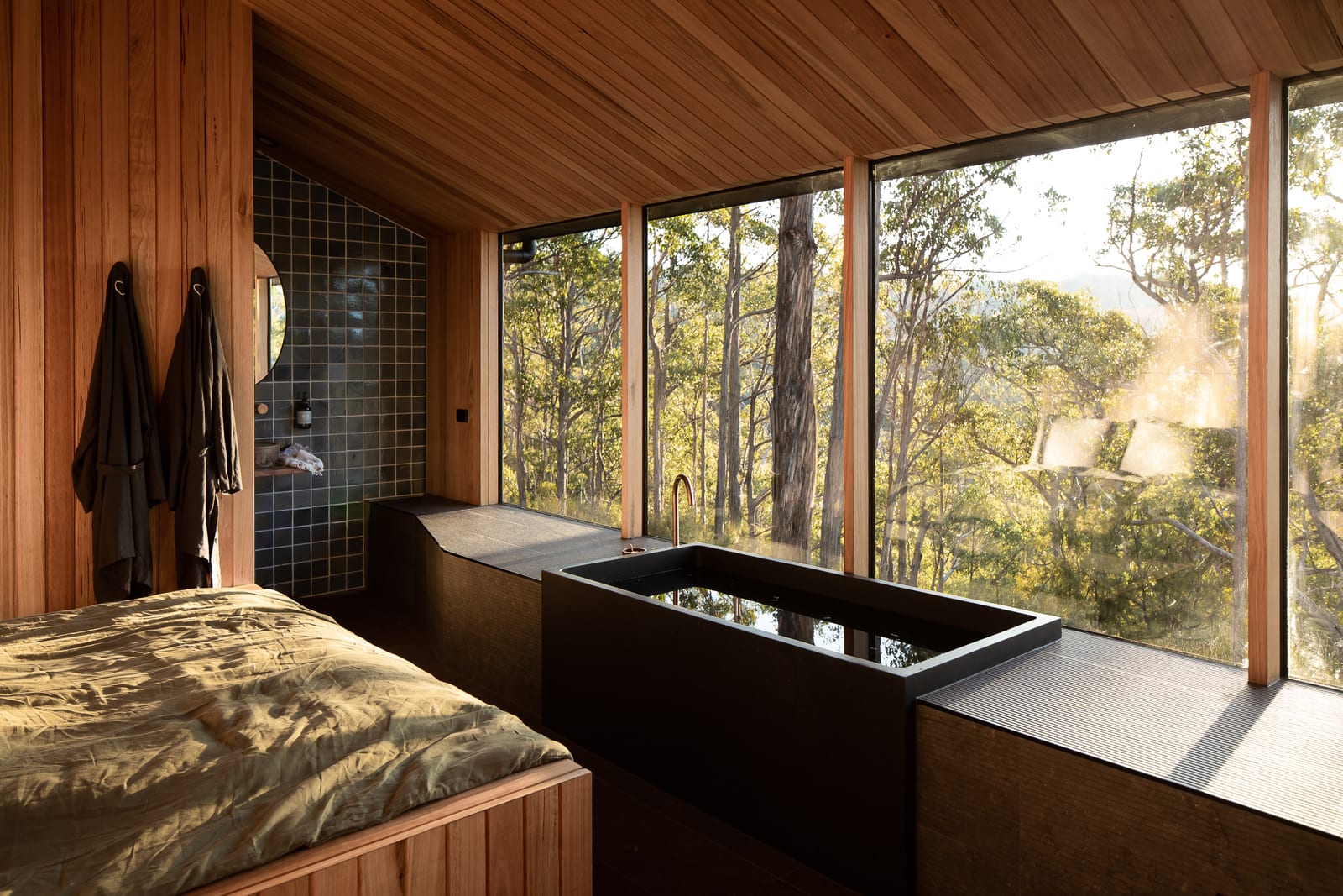 The bed and the bath in one room: olive linen on the left, a black stone bath along a corner of full-height glass, two robes on hooks and the green-tiled ensuite behind, with sunlit gums filling the windows.
