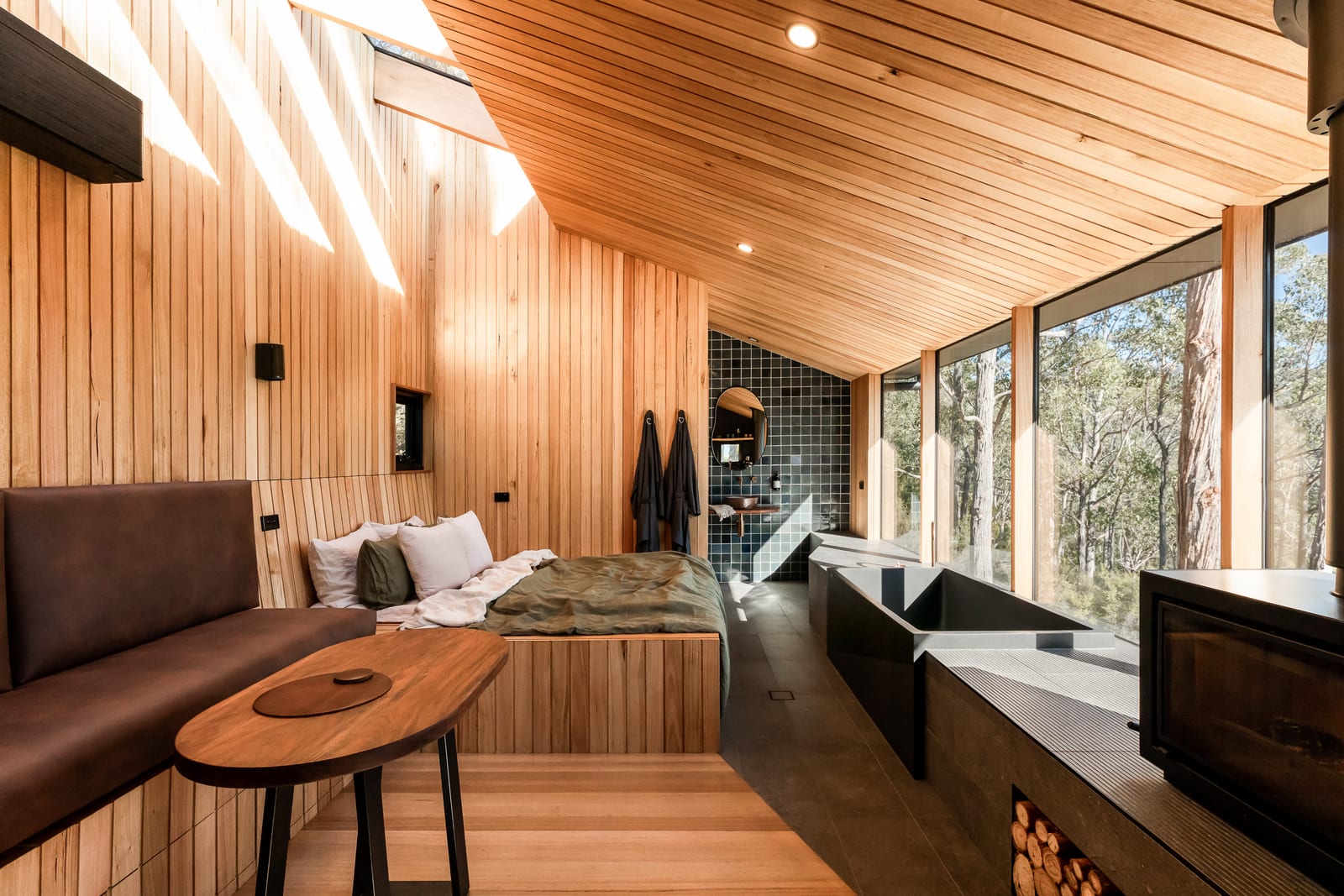 Morning light striking down a timber-lined wall from a long skylight, with a leather bench and round table in the foreground, the bed in olive linen beyond, and the bath and firewood store along the windows to the right.