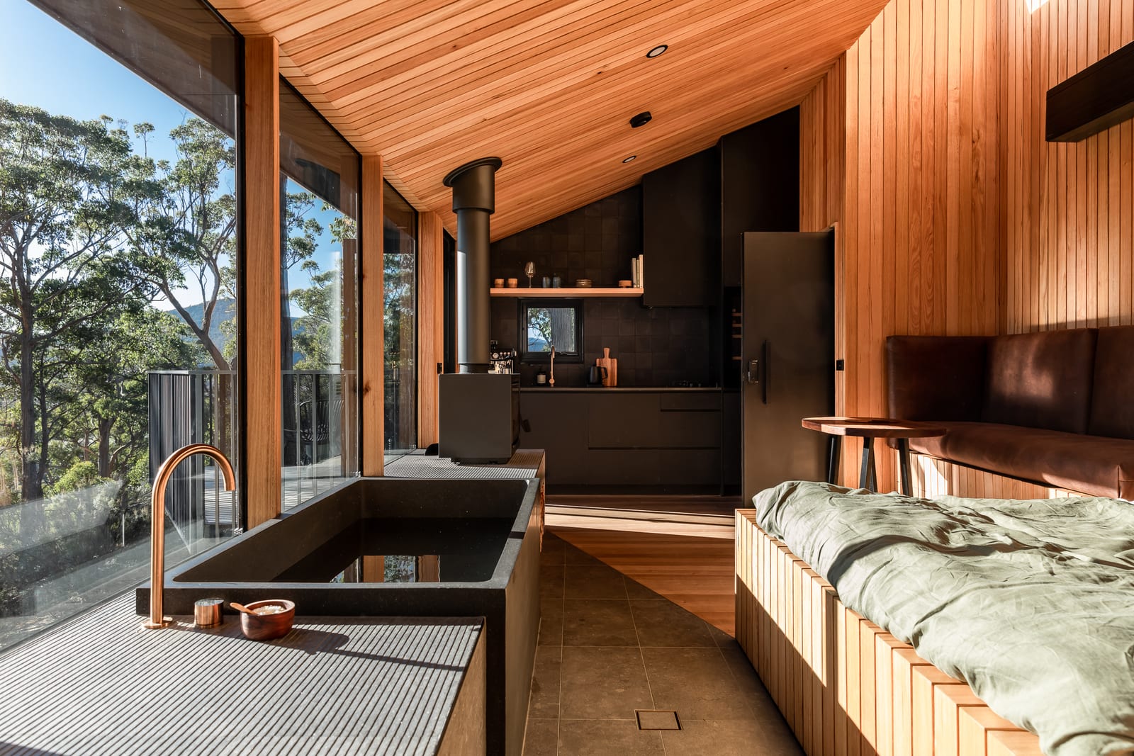 The length of the cabin from the bath end: a copper tap over dark stone, the wood fire and black kitchen in the middle distance, the bed and a leather bench to the right, and glass along the whole left wall opening to the valley.