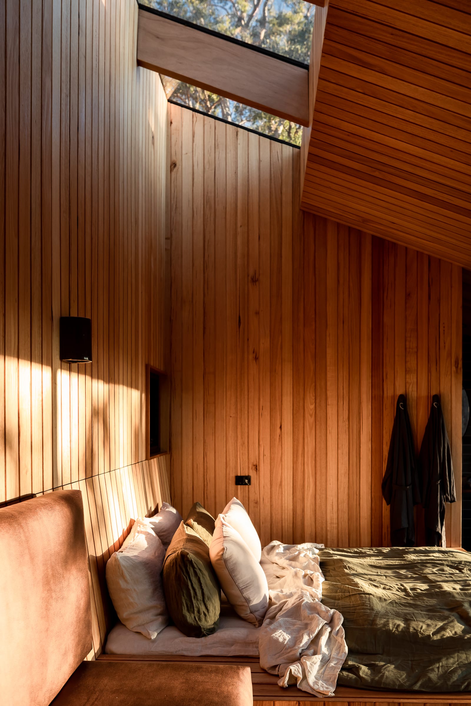 The bedroom of a Hunter cabin: a velvet bench and linen-dressed bed against warm timber walls, lit by a clerestory window