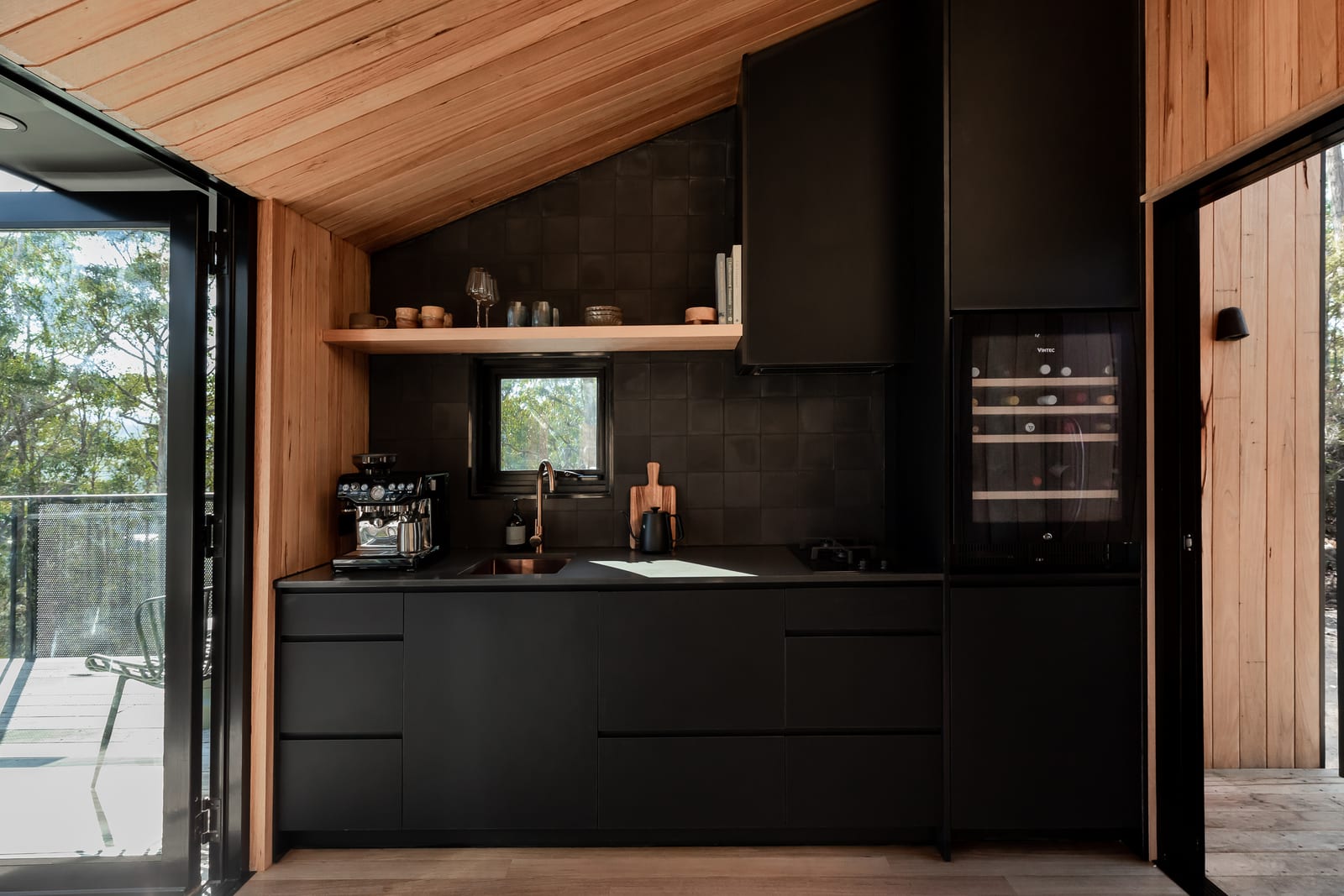 The kitchen: matte black joinery and dark tile under a raked timber ceiling, with an espresso machine, a timber shelf of ceramics and a glass-fronted wine fridge, and the deck door open at the left.