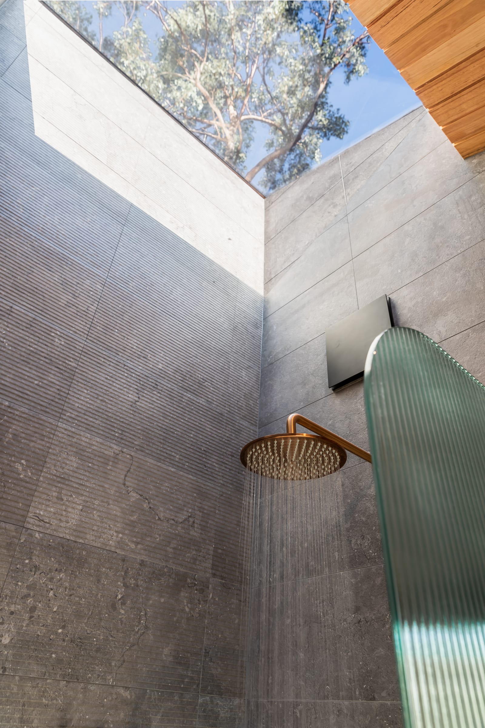 The shower from below: a copper rain head against grey stone walls that open at the top to a skylight, with a gum tree and blue sky directly overhead.