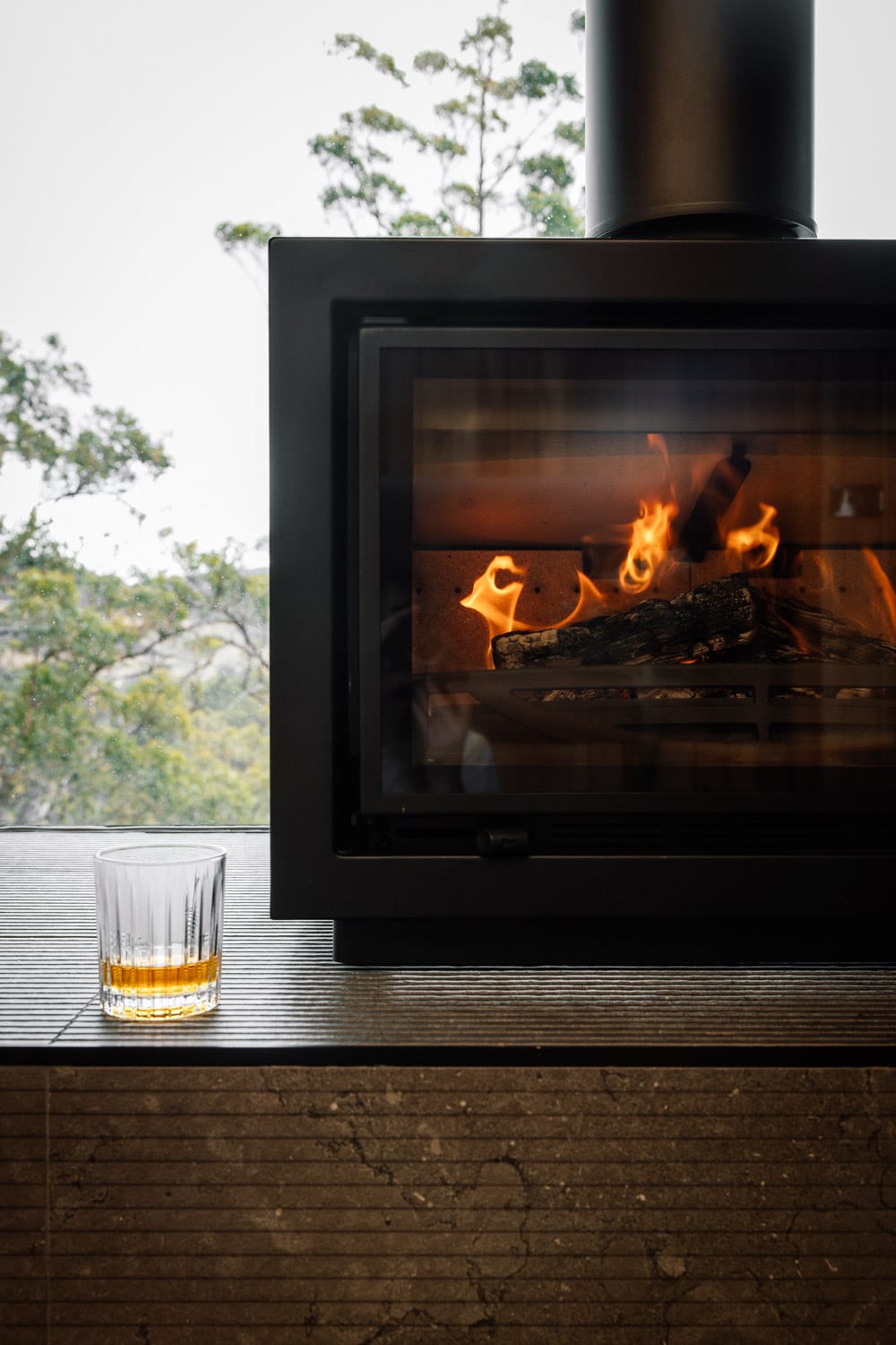 Flames in the cabin's black wood heater, a whisky glass resting on the hearth ledge and bushland through the window beyond