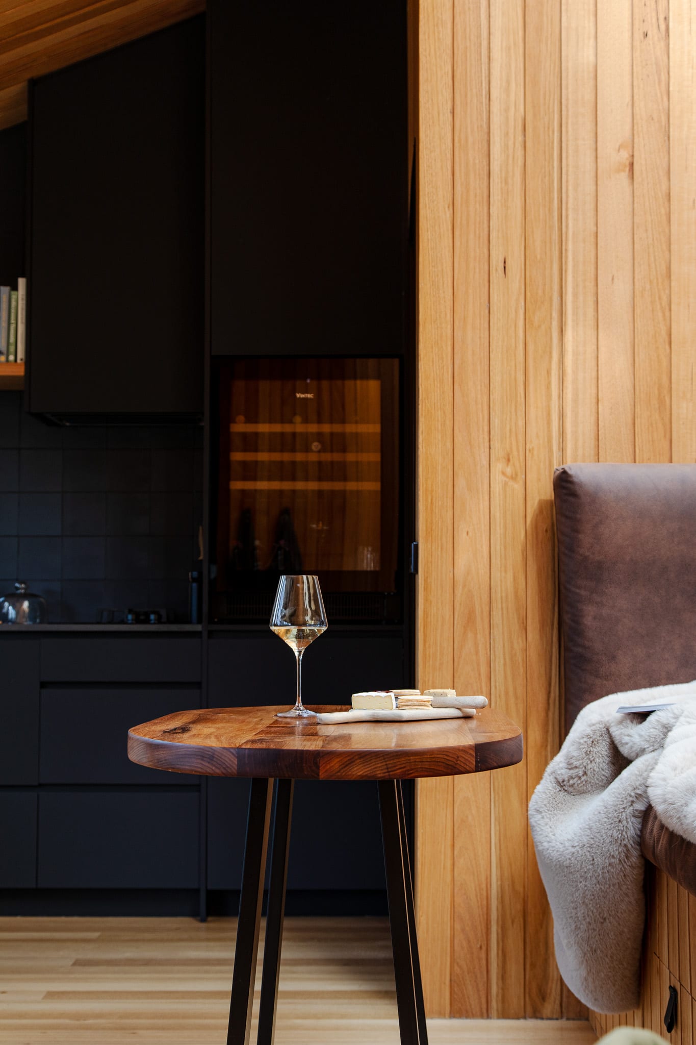 A glass of white wine and a small board set out on a round timber side table inside a Hunter Huon Valley cabin, the dark kitchenette and wine fridge behind