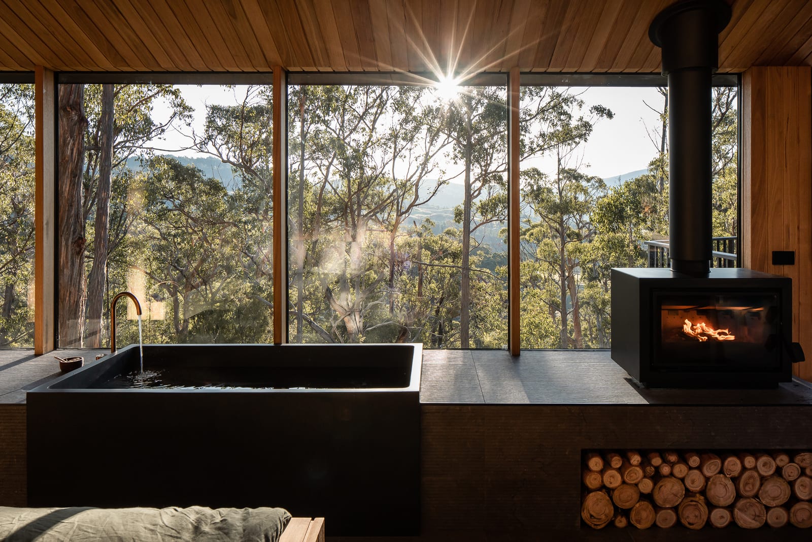 A deep black stone bath beside a lit wood fire inside a Hunter cabin, eucalypts and the valley beyond the full-height windows