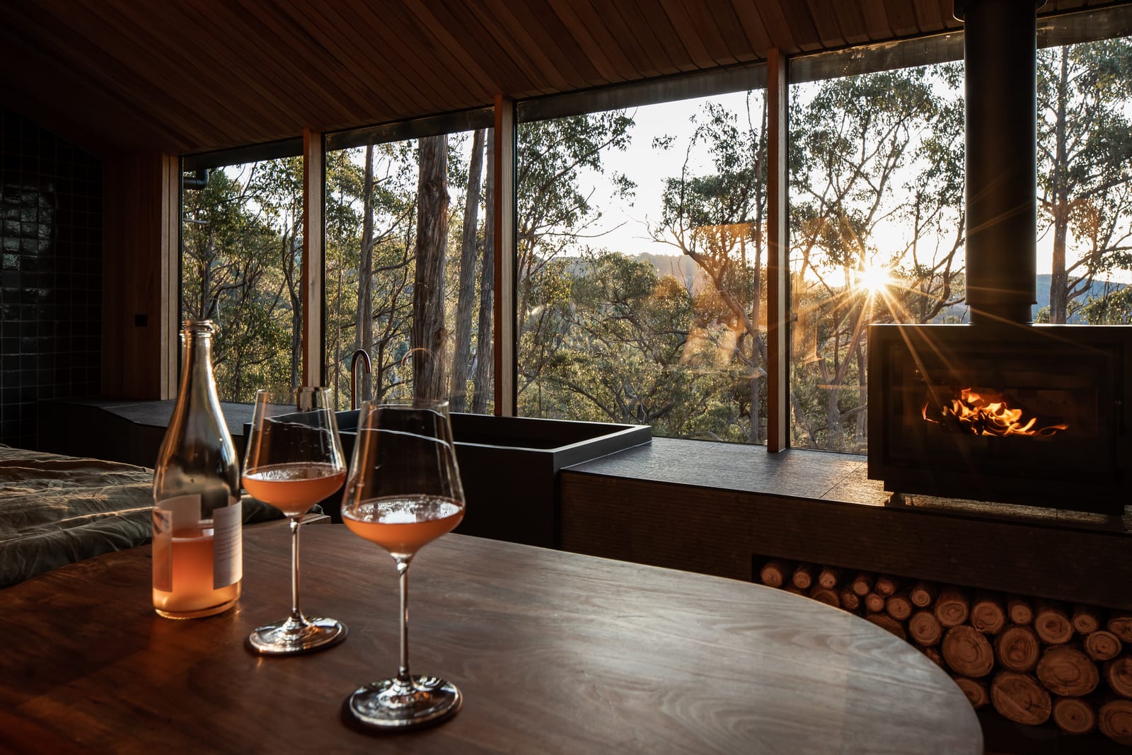Two glasses of sparkling rose on a timber table inside a Hunter cabin, the wood fire alight and the sun setting through the full-height windows behind the stone bath