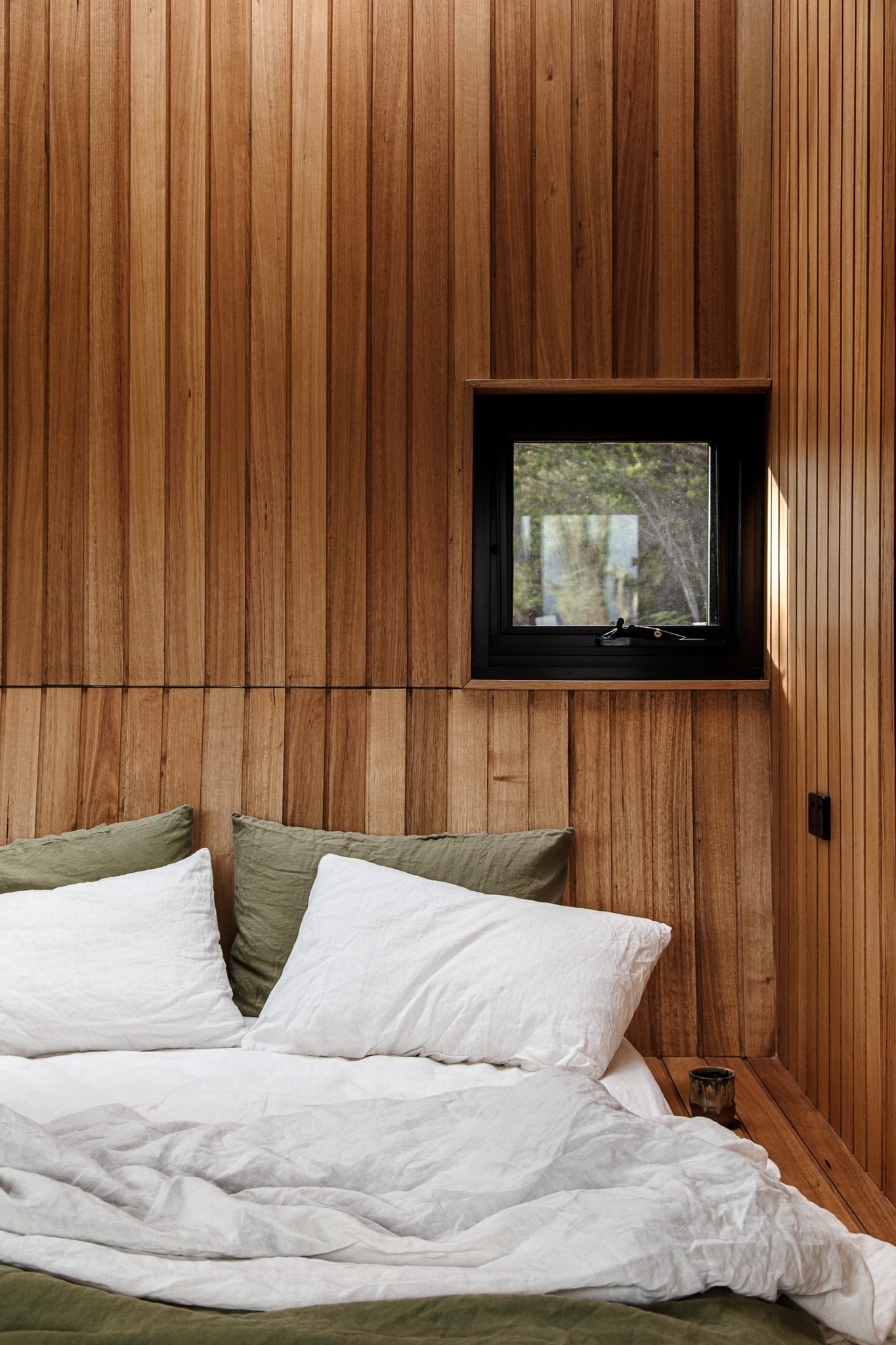 A bed made up in white and olive linen against the timber-lined wall of a Hunter cabin, a small window above it looking out into the bush