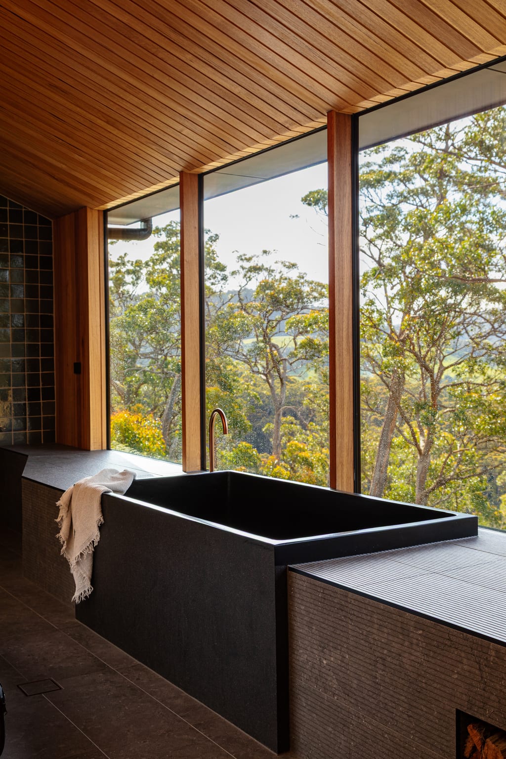 A deep black stone bath set beneath a wall of windows in a Hunter cabin, a linen towel over its edge and gum trees beyond